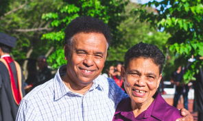 A smiling couple with a student in their graduation gear in the background.