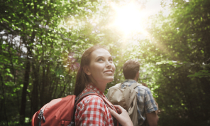 Young woman hiking in a forest and looking up with the sun's glare above her.