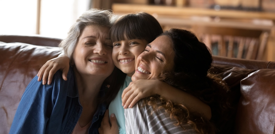 Child hugging her grandmother and mother while sitting with both on a leather sofa.