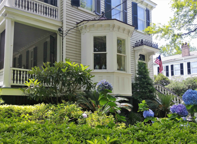 A colonial house with a porch and large window with beautiful green landscaping and hydrangeas in front.
