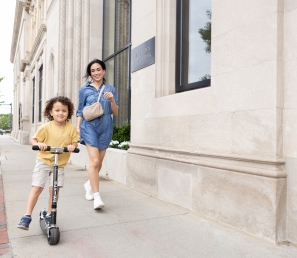 Mom running next to her child on a scooter on a sidewalk by Watertown Savings Bank.