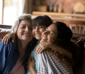 Child hugging her grandmother and mother while sitting with both on a leather sofa.